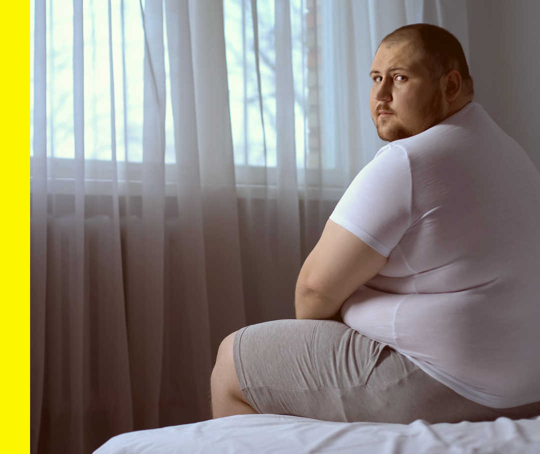 Akne inversa: Überblick Ein übergewichtiger Mensch sitzt auf einem Bett