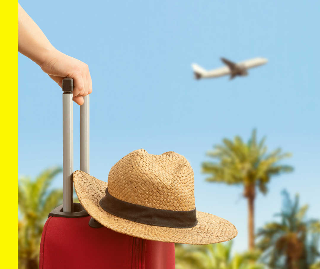 Ab in den Urlaub mit Akne inversa Nahaufnahme eines Huts, der auf einem Rollkoffer liegt. Dahinter Palmen und ein startendes Flugzeug.