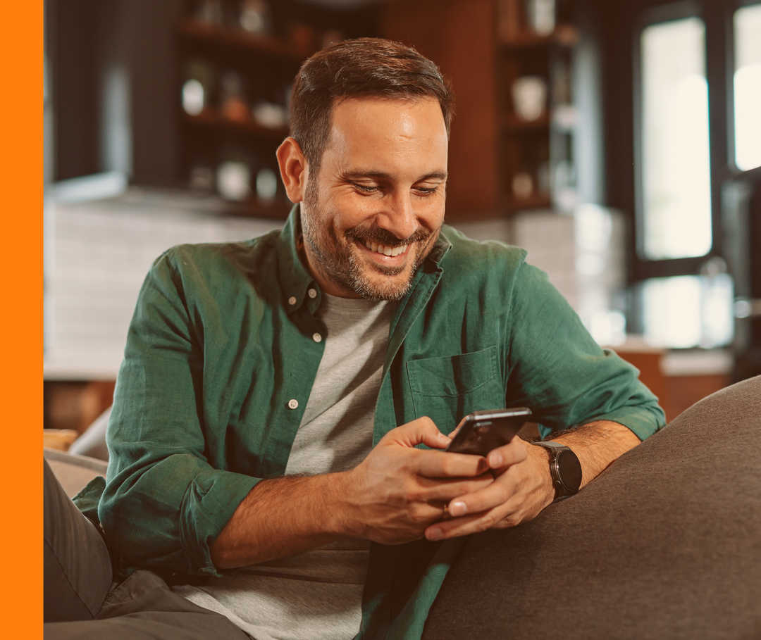 Live Chat des Medizinischen InfoService Eine lächelnde Person sitzt entspannt auf dem Sofa und hält ein Smartphone in der Hand.