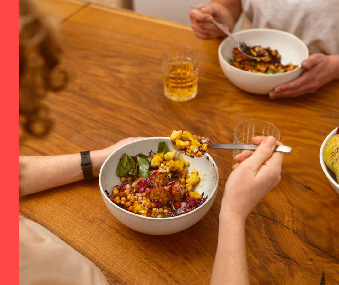 Psoriasis: Ernährung Zwei Personen sitzen an einem Holztisch essen Gerichte aus Schalen.