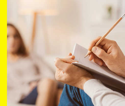 Zwei Personen, von denen eine unscharf im Hintergrund zu sehen ist, während die andere etwas in ein Notizbuch schreibt. 