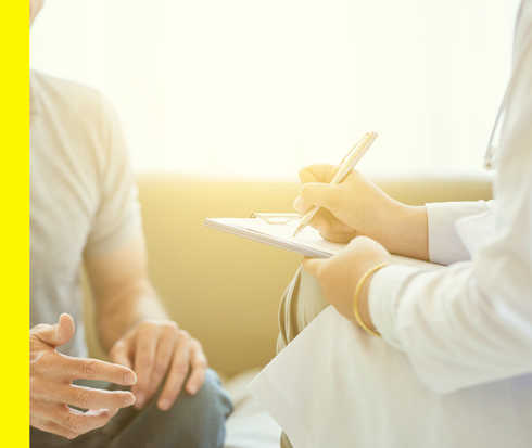 Bildausschnitt von zwei Personen, deren Gesichter nicht zu sehen sind, im Gespräch. Die eine trägt einen weißen Kittel und macht sich Notizen.