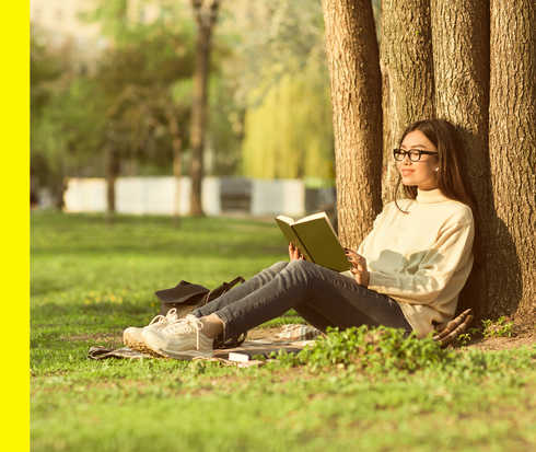Eine Frau mit Brille lehnt sitzend an einem Baum und liest lächelnd ein Buch.