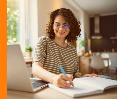 Eine lächelnde Person sitzt vor einem aufgeklappten Laptop und schreibt in ein Notizbuch. 