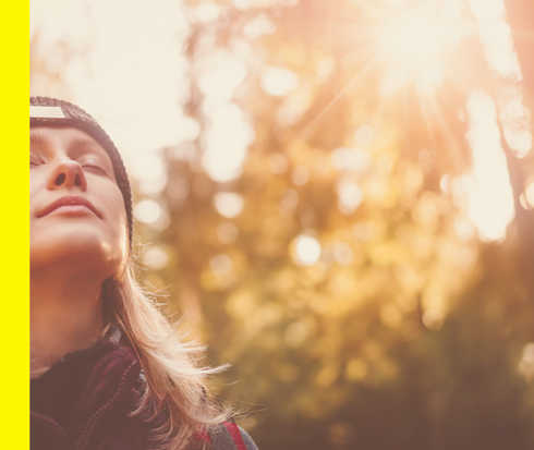 Eine Frau steht vor sonnendurchfluteten Bäumen und blickt mit geschlossenen Augen nach oben.