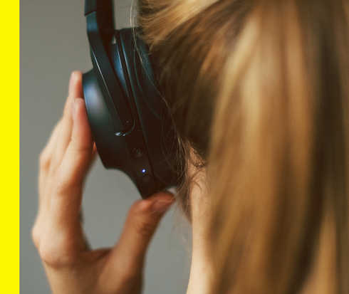 Der Hinterkopf einer Frau, die einen Kopfhörer trägt, den sie mit einer Hand fixiert.