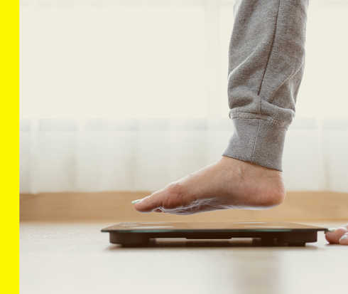 Bildausschnitt mit zwei Füßen, von denen einer über einer Waage schwebt.