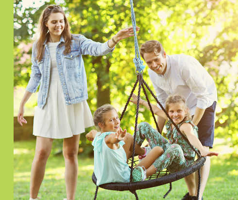 Eine Frau und ein Mann spielen mit zwei Kindern, die in einer Schaukel sitzen, im Freien.