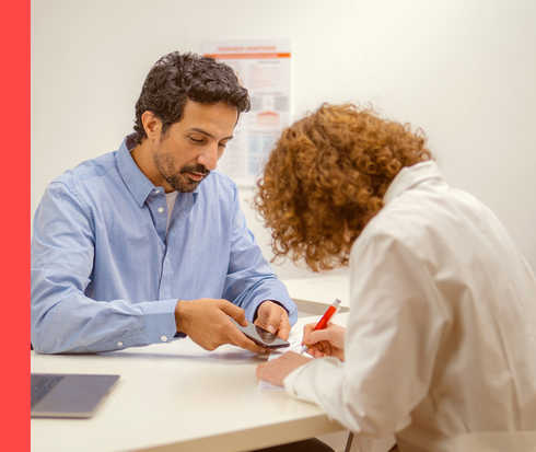 Ein Mann in einem blauen Hemd zeigt einer Frau in einem weißen Kittel sein Smartphone. Sie macht sich Notizen.