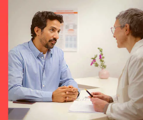 Ein bärtiger Mann in einem blauen Hemd und eine Frau in einem weißen Kittel sitzen an einem Tisch und unterhalten sich.