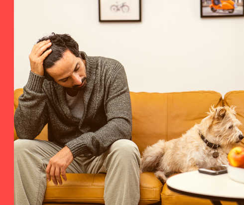 Ein Mann sitzt mit erschöpftem Gesichtsausdruck auf einem Sofa und hält sich mit der rechten Hand den Kopf. Neben ihm liegt ein Hund.