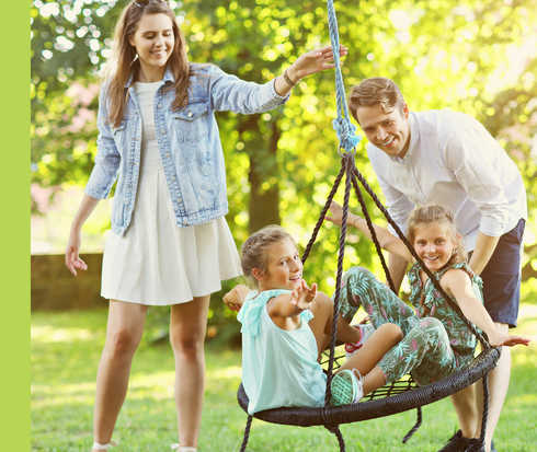 Eine Frau und ein Mann spielen mit zwei Kindern, die in einer Schaukel sitzen, im Freien.