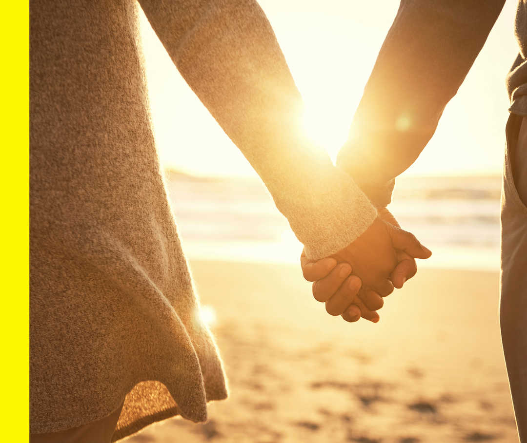 Nahaufnahme von zwei Personen, die Hand in Hand am Strand im goldenen Licht stehen.
