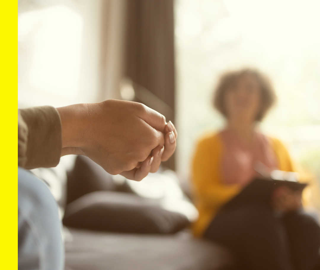 Im Vordergrund ineinander gelegte Hände und im Hintergrund eine unscharfe Person, die ein Klemmbrett hält