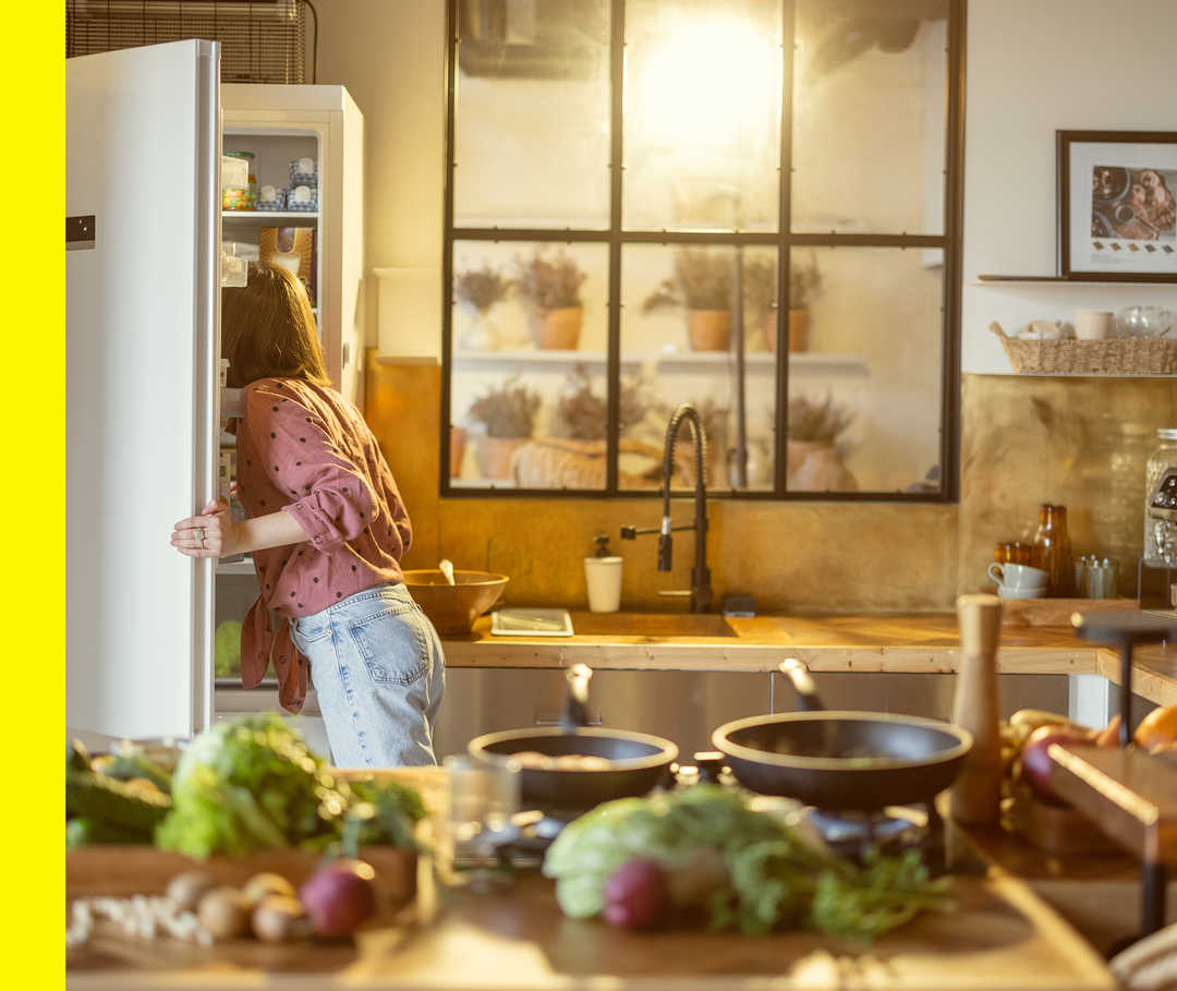 Eine Frau sucht im Hintergrund Zutaten aus einem Kühlschrank, während im Vordergrund schon viele Zutaten liegen.