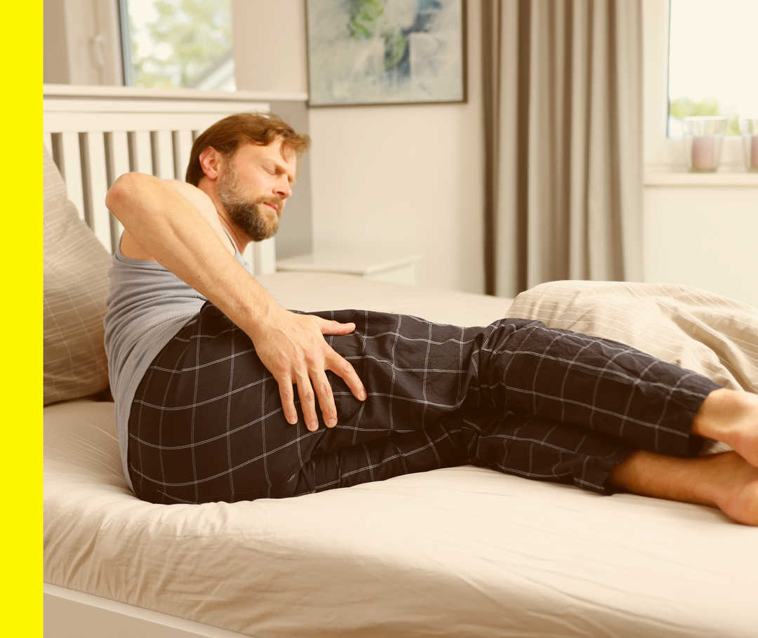 Ein Mann liegt im Bett und fasst sich mit schmerzverzerrtem Gesicht an das Gesäß.