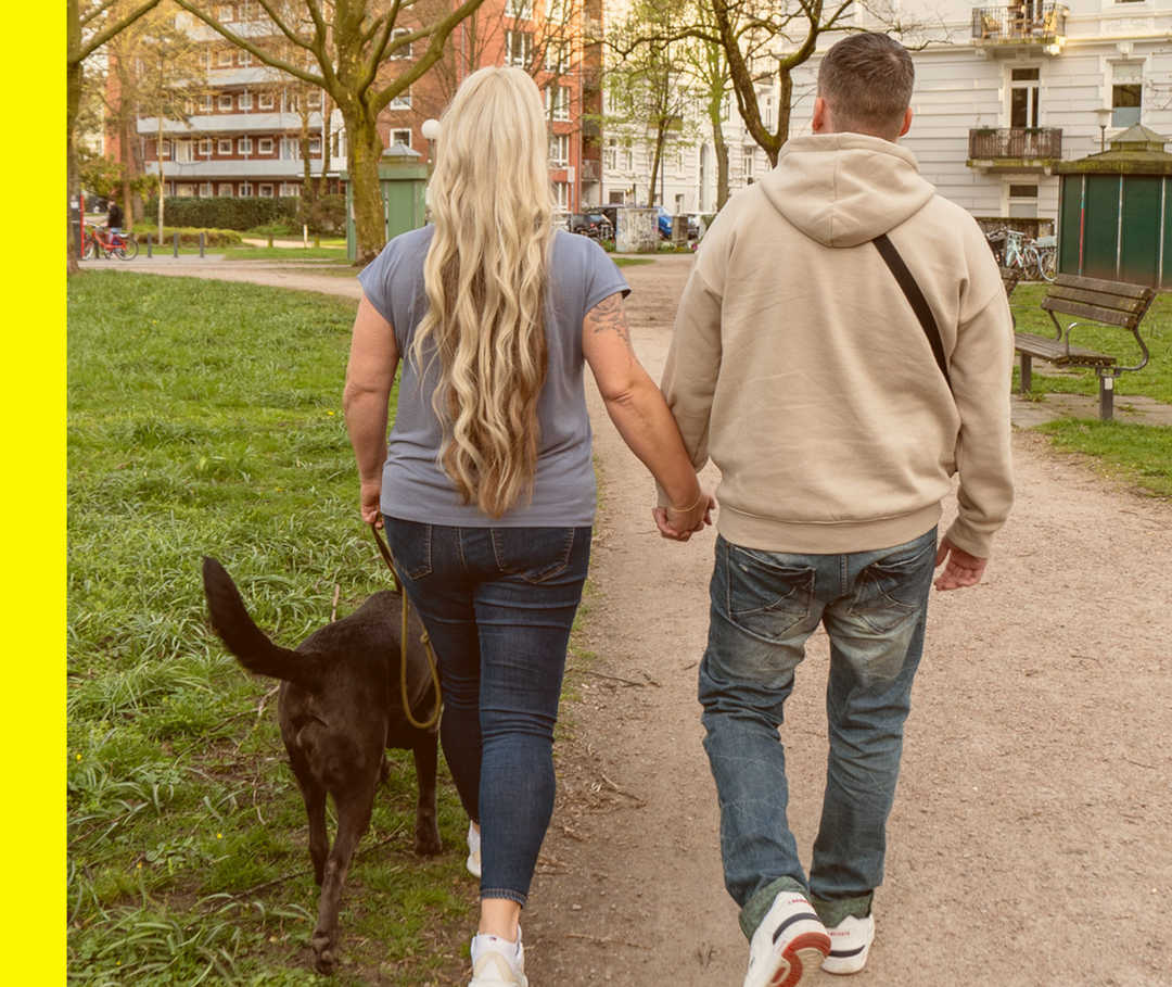 Rabea und ihr Freund gehen händchenhaltend mit ihrer Hündin Berta in einem Park spazieren.