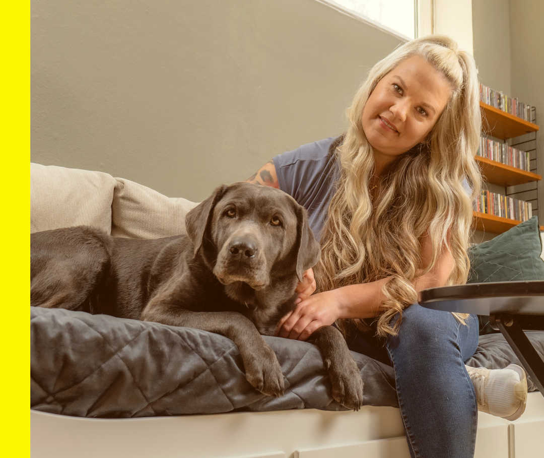 Rabea sitzt mit ihrer Labradorhündin Berta auf einem Sofa und schaut in die Kamera.