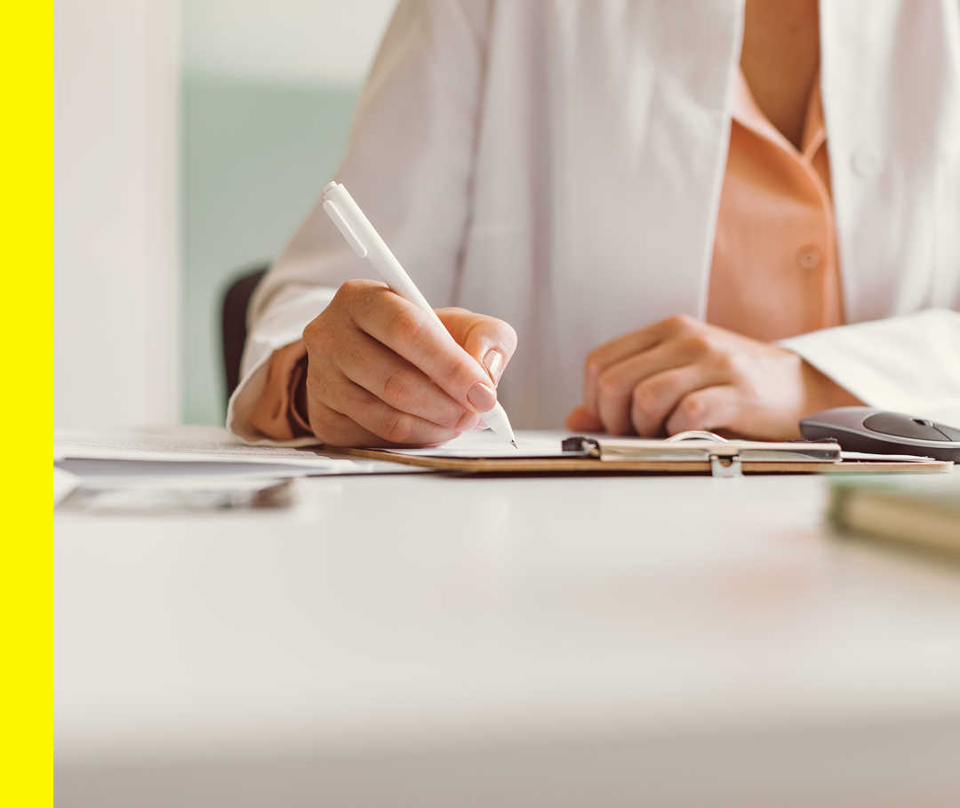 Nahaufnahme einer Person im weißen Kittel, die an einem Schreibtisch sitzt und einen Stift hält.