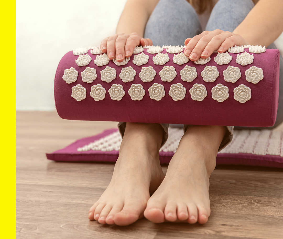 Eine Person sitzt auf einer Akupressurmatte und hält eine weitere aufgerollte Matte fest.