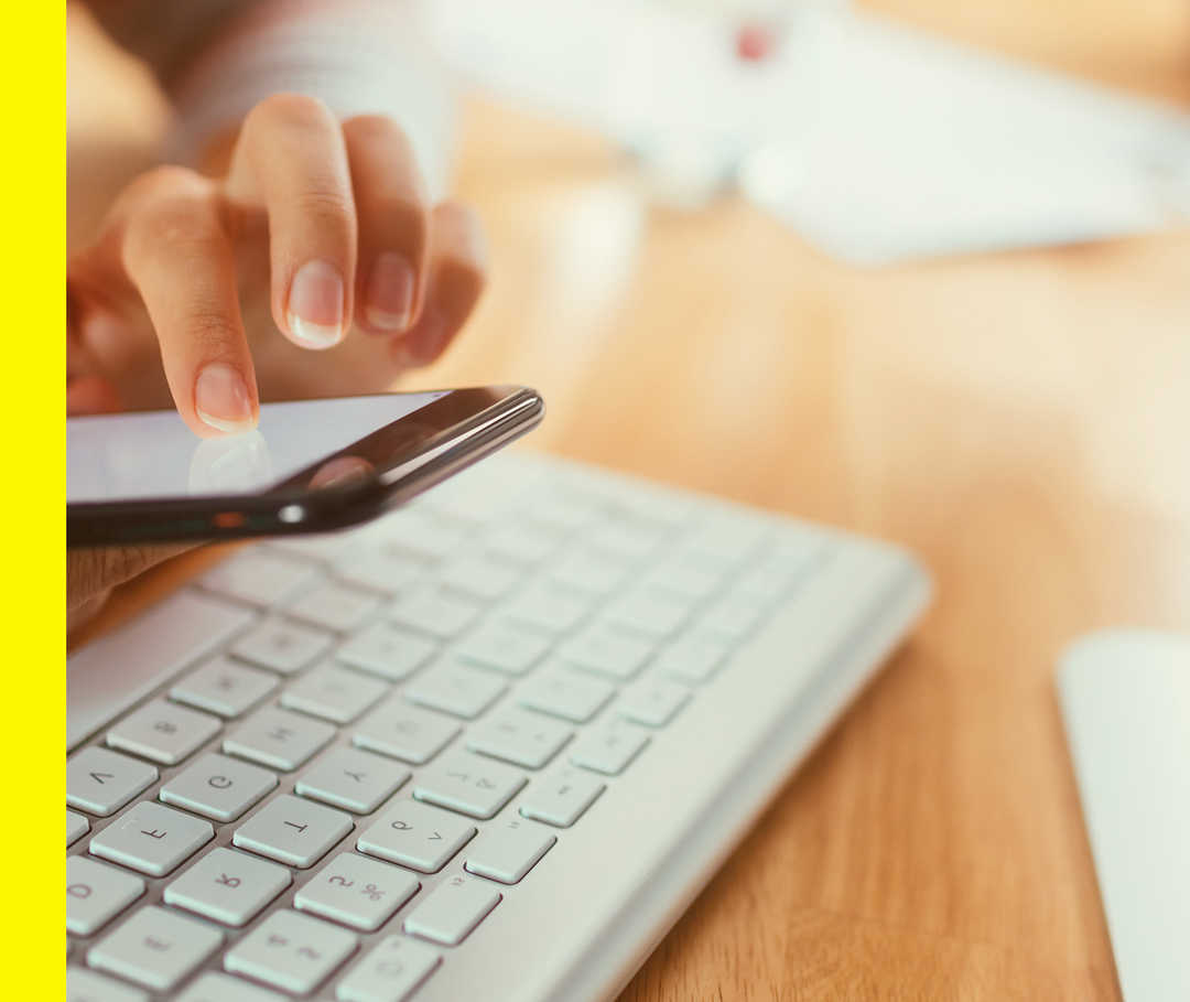 Nahaufnahme von zwei Händen über einer Computertastatur, die ein Handy halten. Ein Finger berührt den leuchtenden Bildschirm.