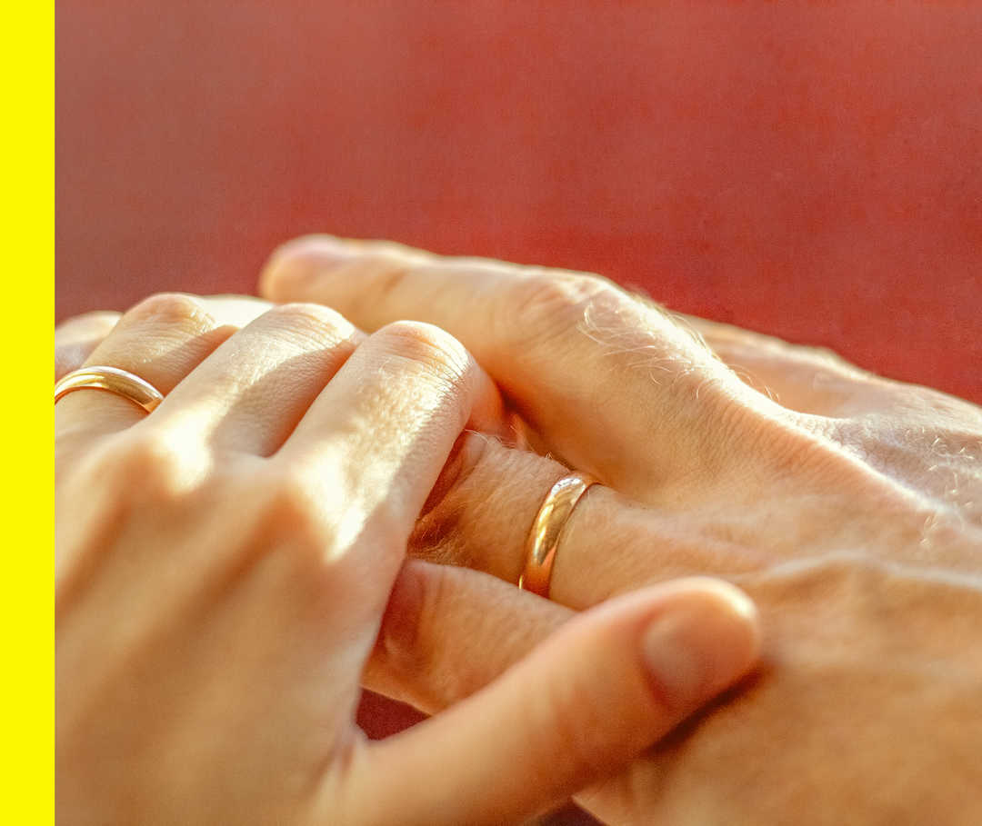 Die Hand einer Person berührt die Hand einer anderen Person. Beide tragen einen Ring am Ringfinger.