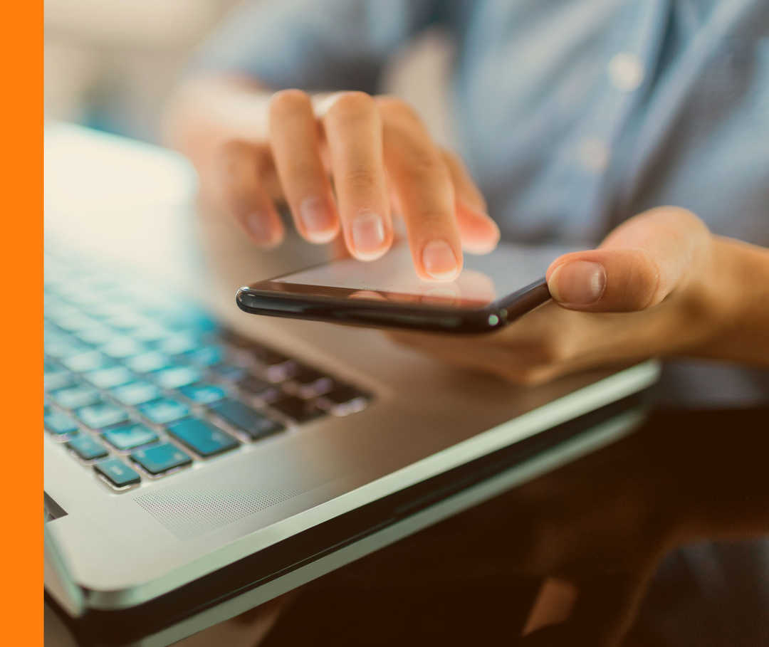 Nahaufnahme einer Person, die vor einem Laptop sitzt und ein Smartphone in der Hand hält.