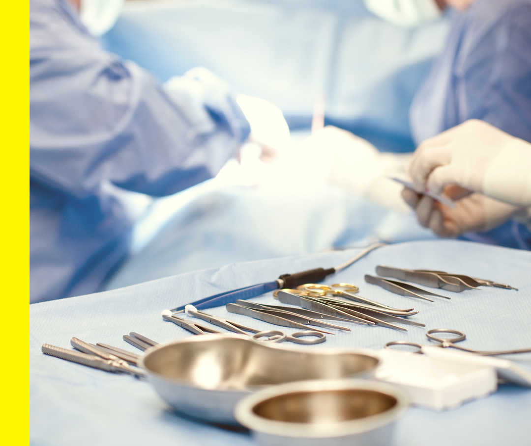 Nahaufnahme des ausgebreiteten Operationsbestecks. Im Hintergrund sind zwei Personen mit Handschuhen und blauen Kitteln.