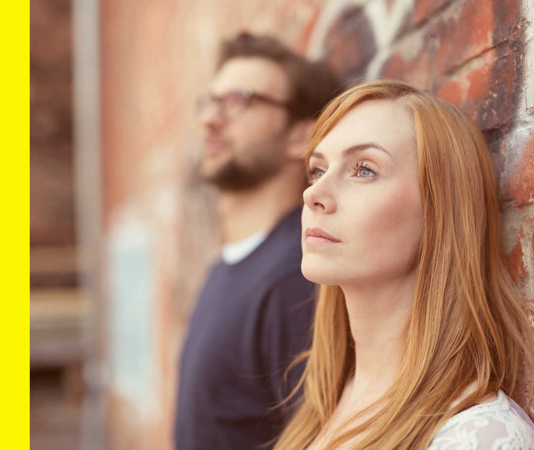 Eine blonde Frau und ein Mann mit Bart und Brille lehnen an einer Ziegelsteinmauer