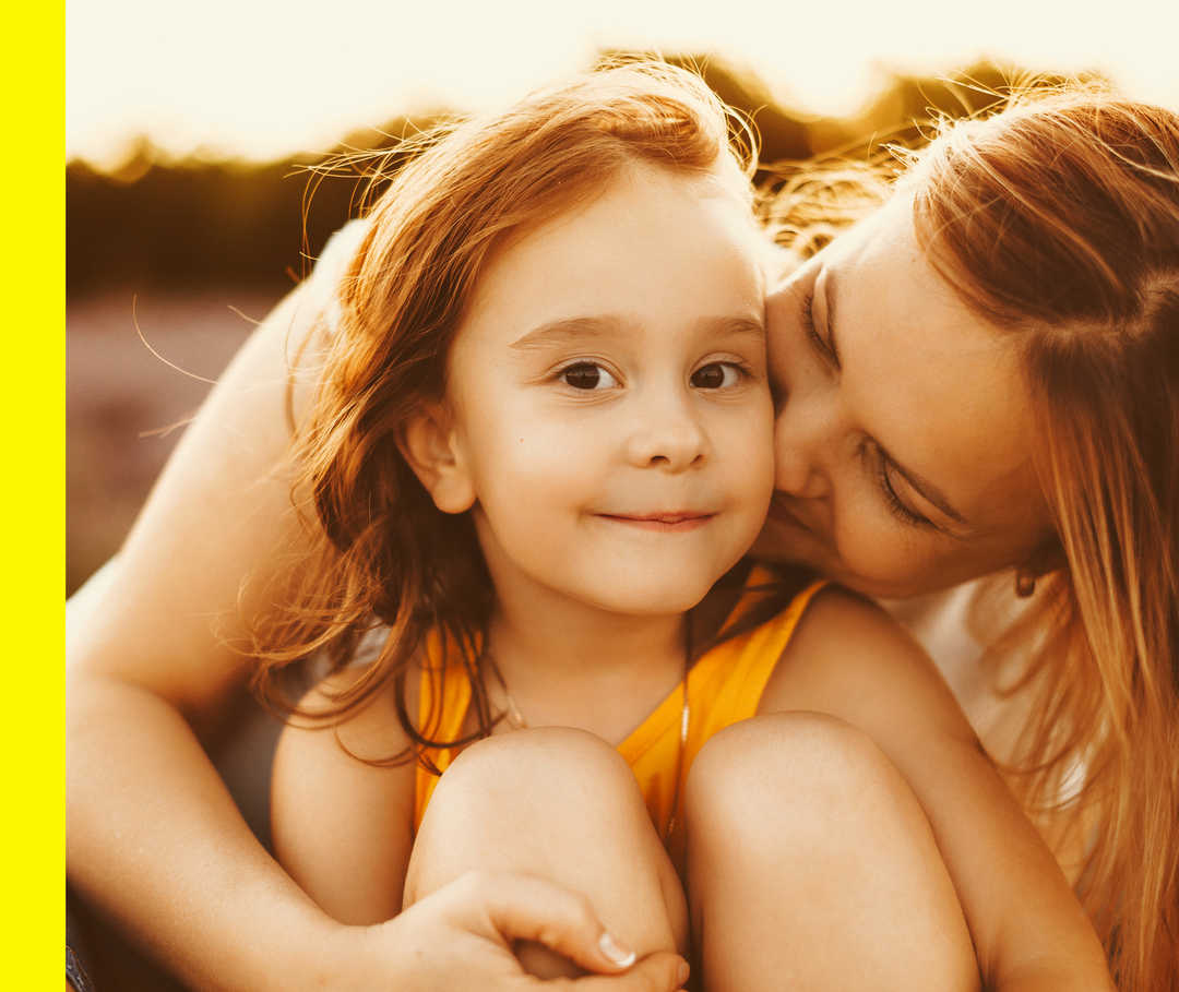 Eine Mutter gibt ihrer Tochter einen Kuss die in die Kamera lächelt.