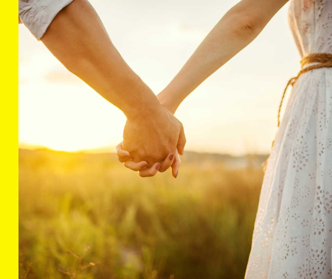 Ein Pärchen hält sich an den Händen fest, im Hintergrund ist ein Sonnenuntergang zu sehen.
