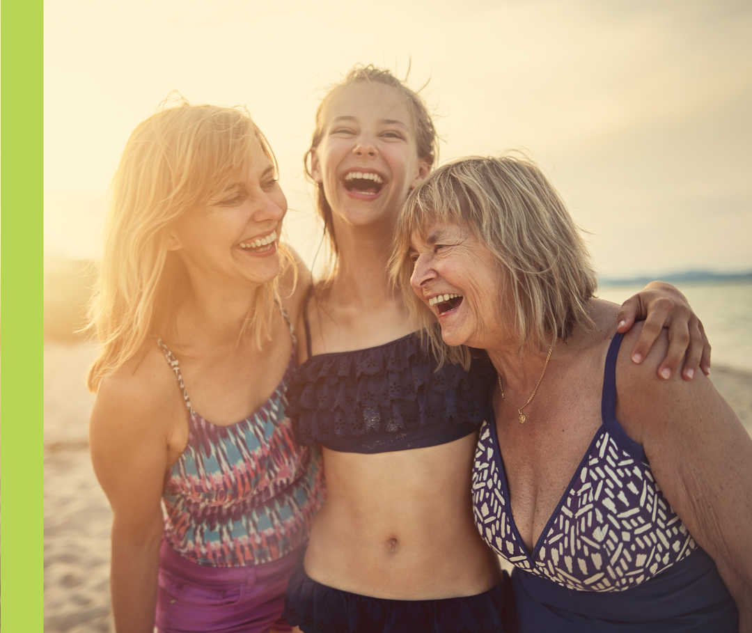 Drei Frauen unterschiedlichen Alters in Strandbekleidung umarmen sich lachend. 