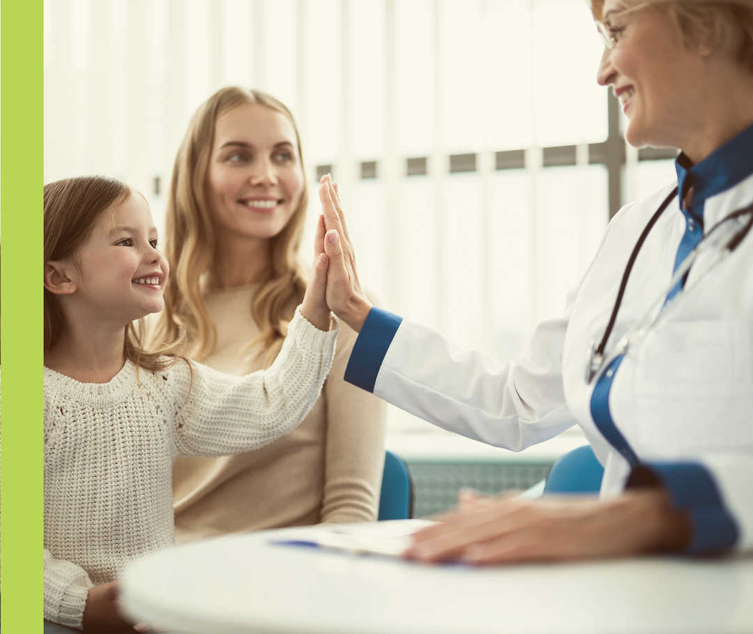 Ein Mädchen klatscht sich mit einer Frau im weißen Kittel ab. Eine zweite Frau schaut lächelnd zu. 