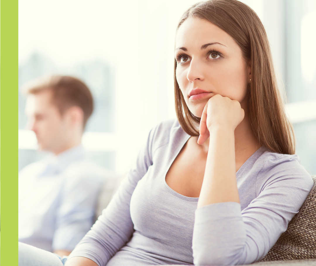 Eine Frau mit nachdenklichem Blick sitzt mit einigem Abstand neben einem Mann, der nur unscharf zu erkennen ist.