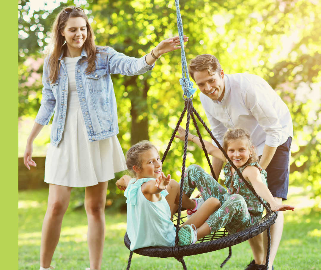 Eine Frau und ein Mann spielen mit zwei Kindern, die in einer Schaukel sitzen, im Freien.