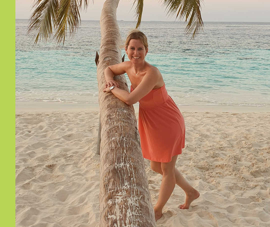 Sandy stützt sich am Strand an einer Palme ab und lächelt in die Kamera.