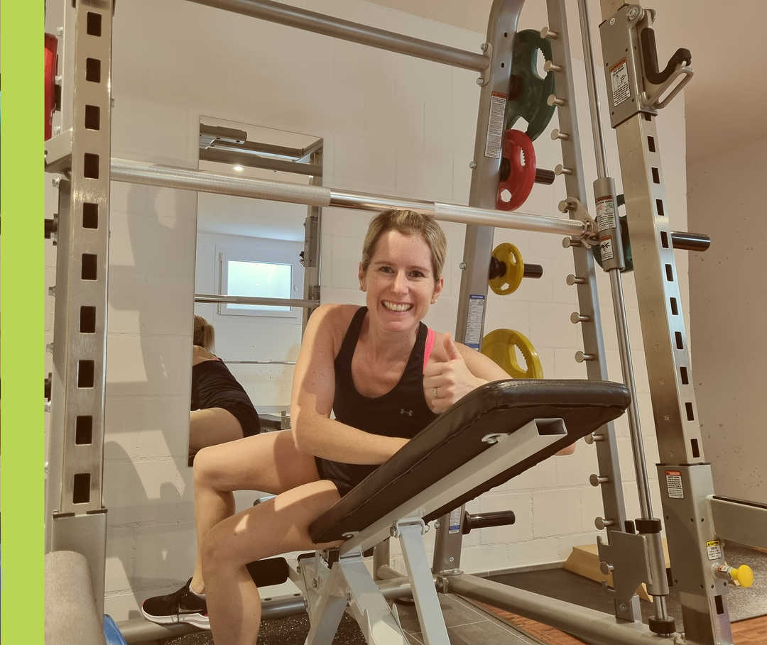 Sandy lacht in Sportkleidung von einer Hantelbank in die Kamera und hält ihren Daumen hoch.
