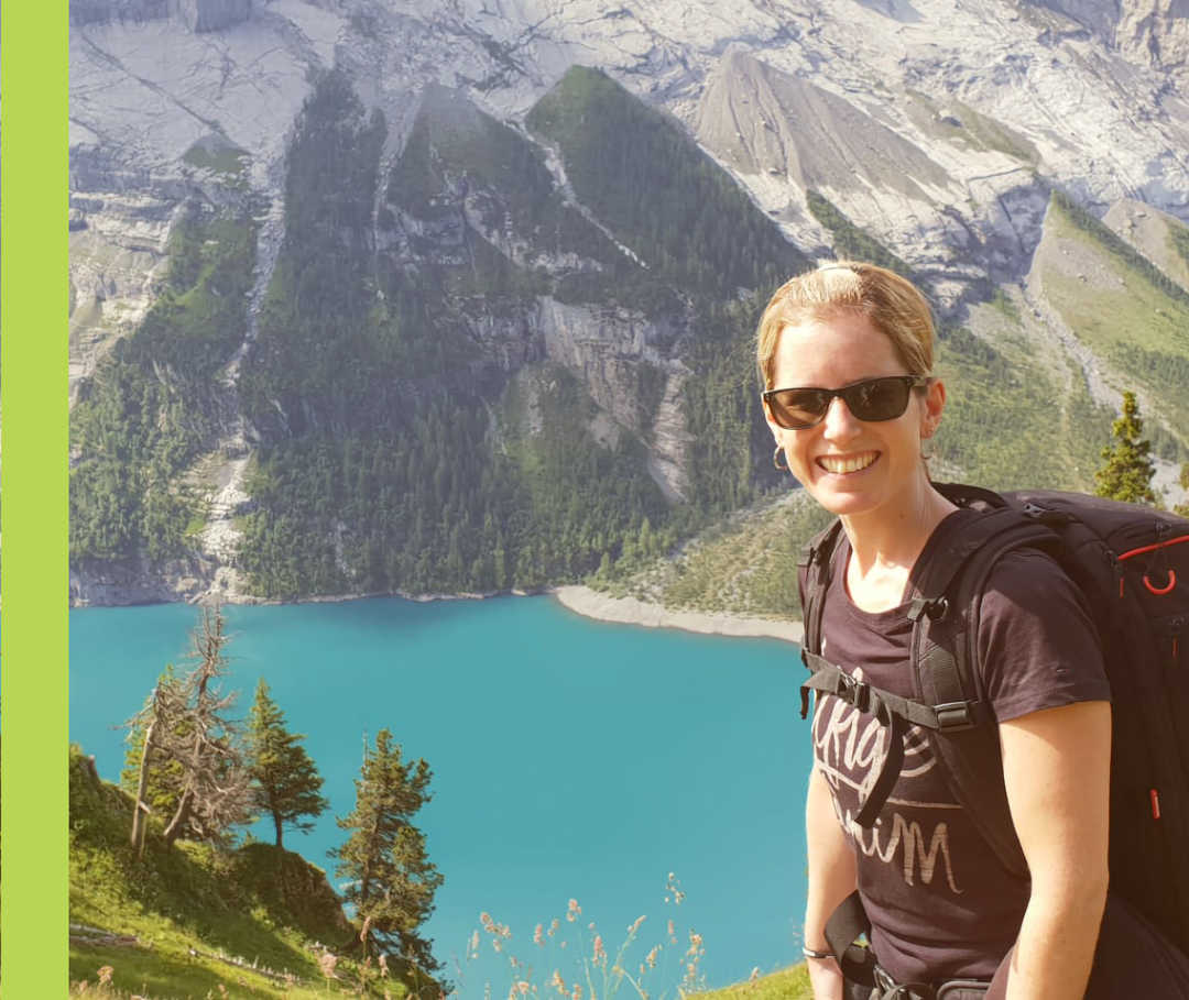 Sandy lächelt vor einem Bergpanorama mit See in die Kamera.