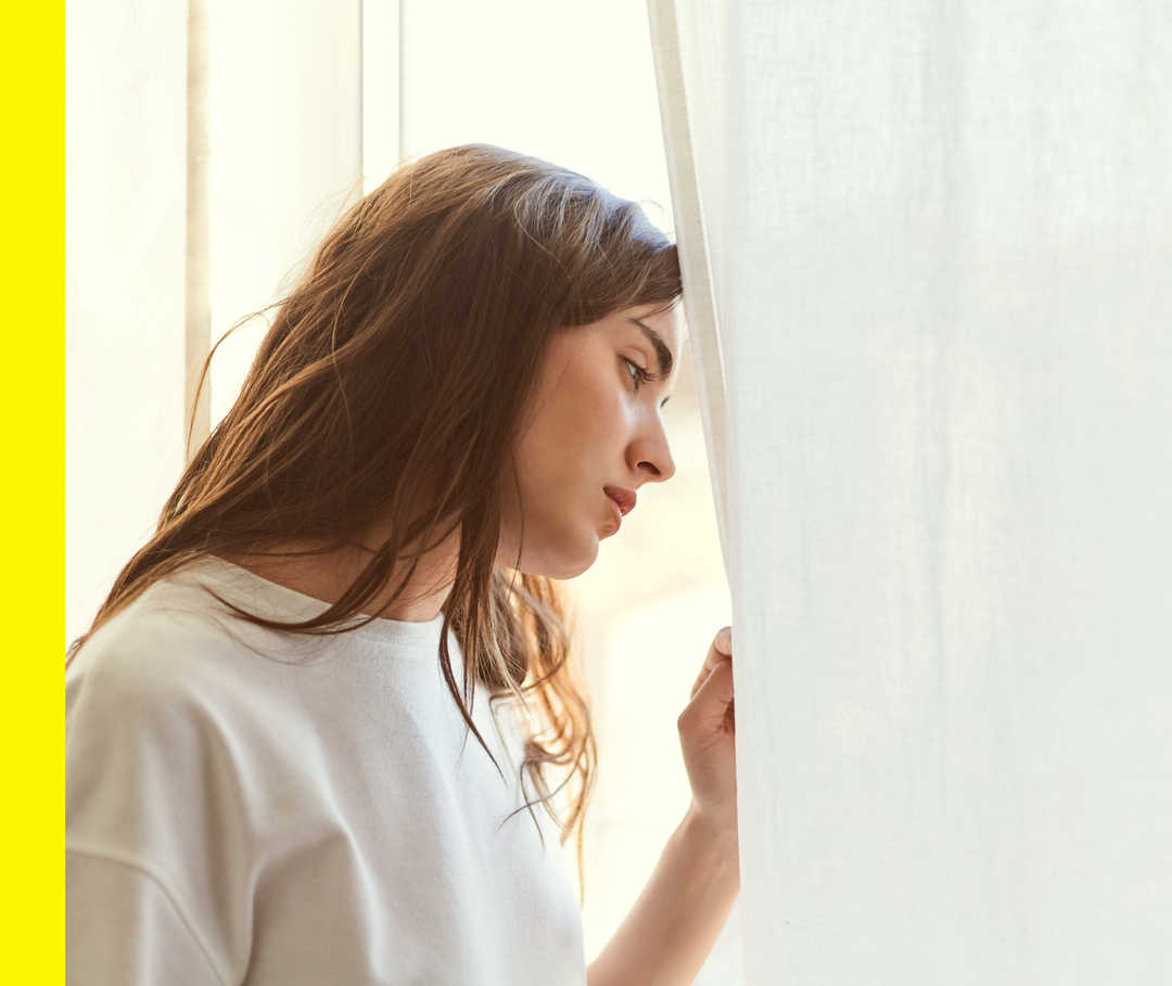 Eine junge Frau steht am Fenster und lehnt ihren Kopf gegen die Scheibe.