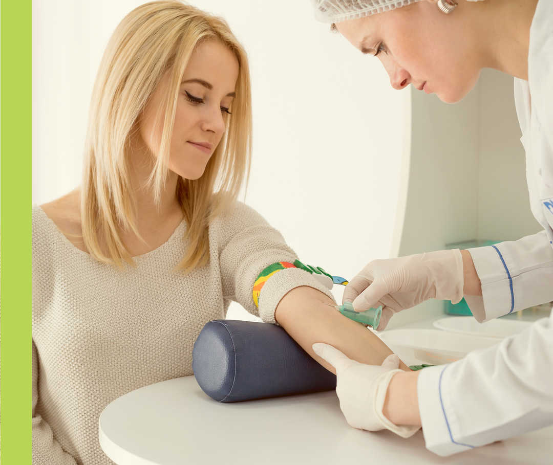 Eine Frau mit Handschuhen und Haarschutz nimmt einer anderen Frau in der linken Armbeuge Blut ab.