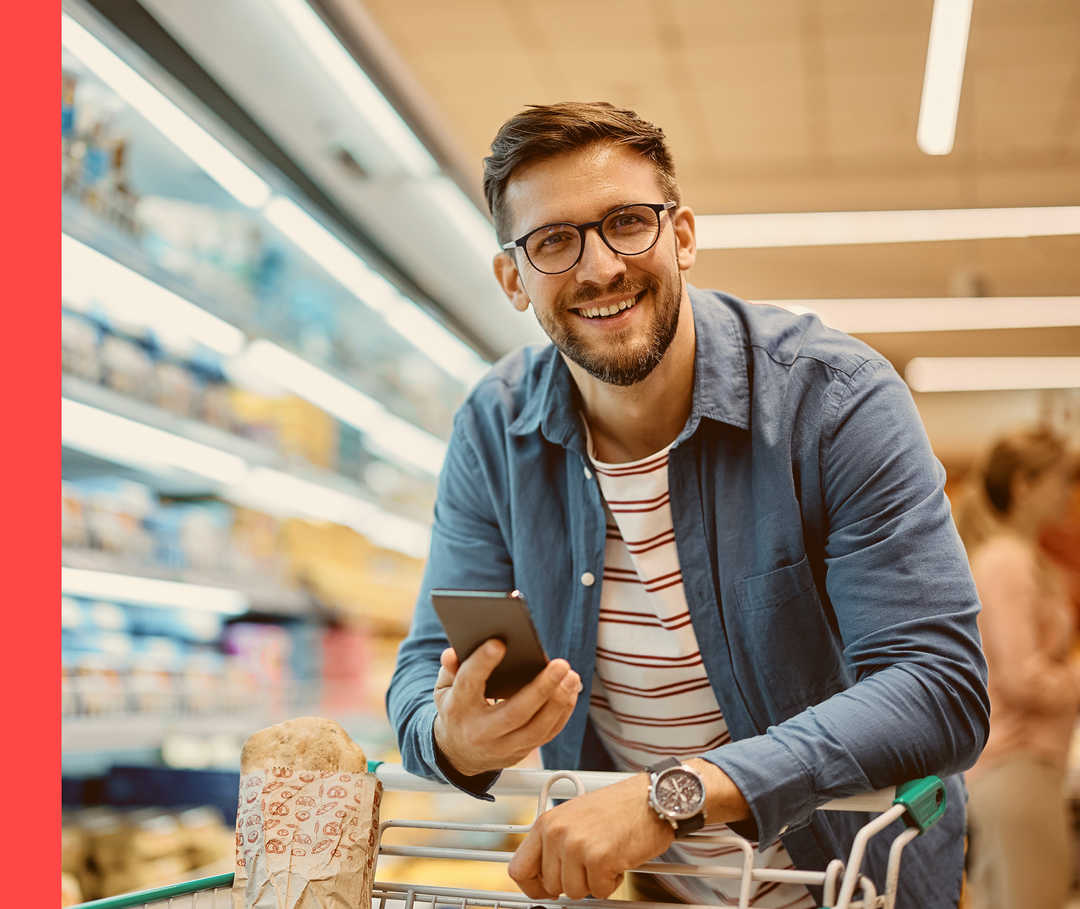 Männliche Person steht mit Smartphone und gefülltem Einkaufswagen vor einem Supermarktregal.