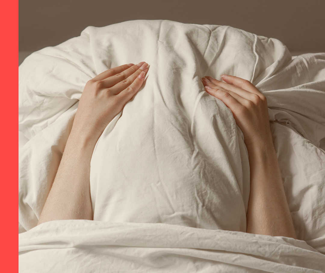 Nahaufnahme von einer Person im Bett, die mit einem Kissen ihr Gesicht verdeckt.