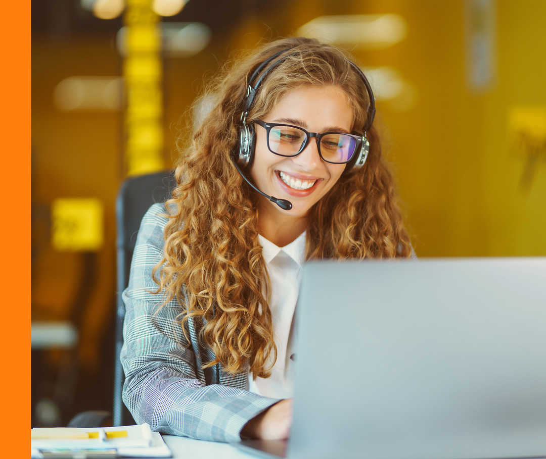 Eine lächelnde Person sitzt mit Headset an einem Schreibtisch und schaut auf ihr geöffnetes Laptop.