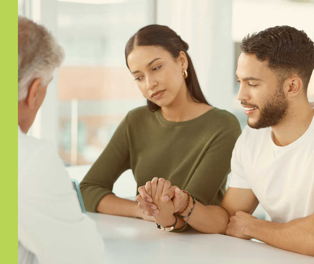 Zwei händchenhaltende Personen sitzen an einem Tisch vor einer Person in einem weißen Kittel. 