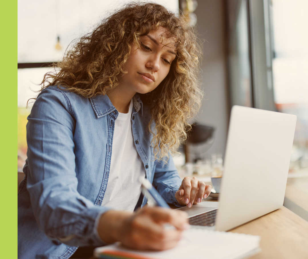 Eine Frau sitzt vor einem Laptop und schreibt dabei auf einen Block.