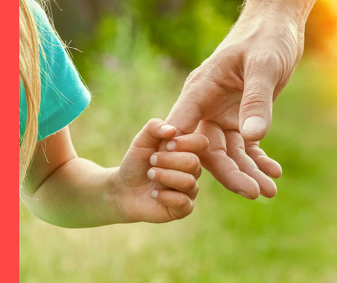 Ein Kind hält den Finger der Hand eines Erwachsenen fest. Im Hintergrund ist unscharf abgebildete Natur.