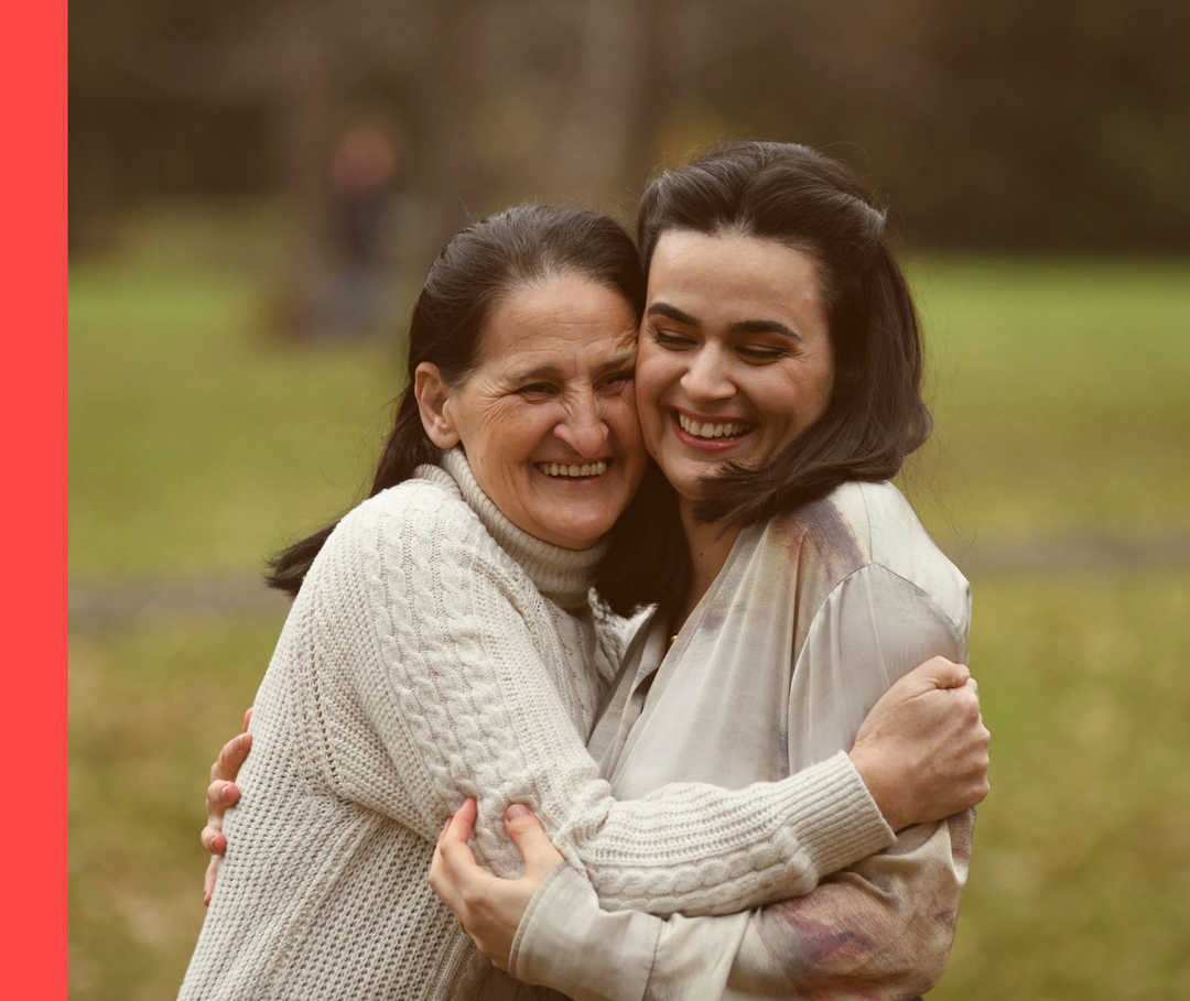 Sine und ihre Mutter stehen im Freien und umarmen sich lachend.
