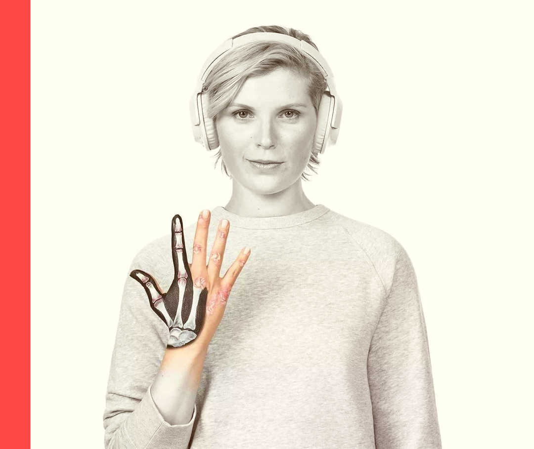 Psoriasis: Patientengeschichten Eine Frau, die Kopfhörer trägt, hält ihre rechte Hand hoch, in der symbolisch mehrere Fingerknochen zu erkennen sind.