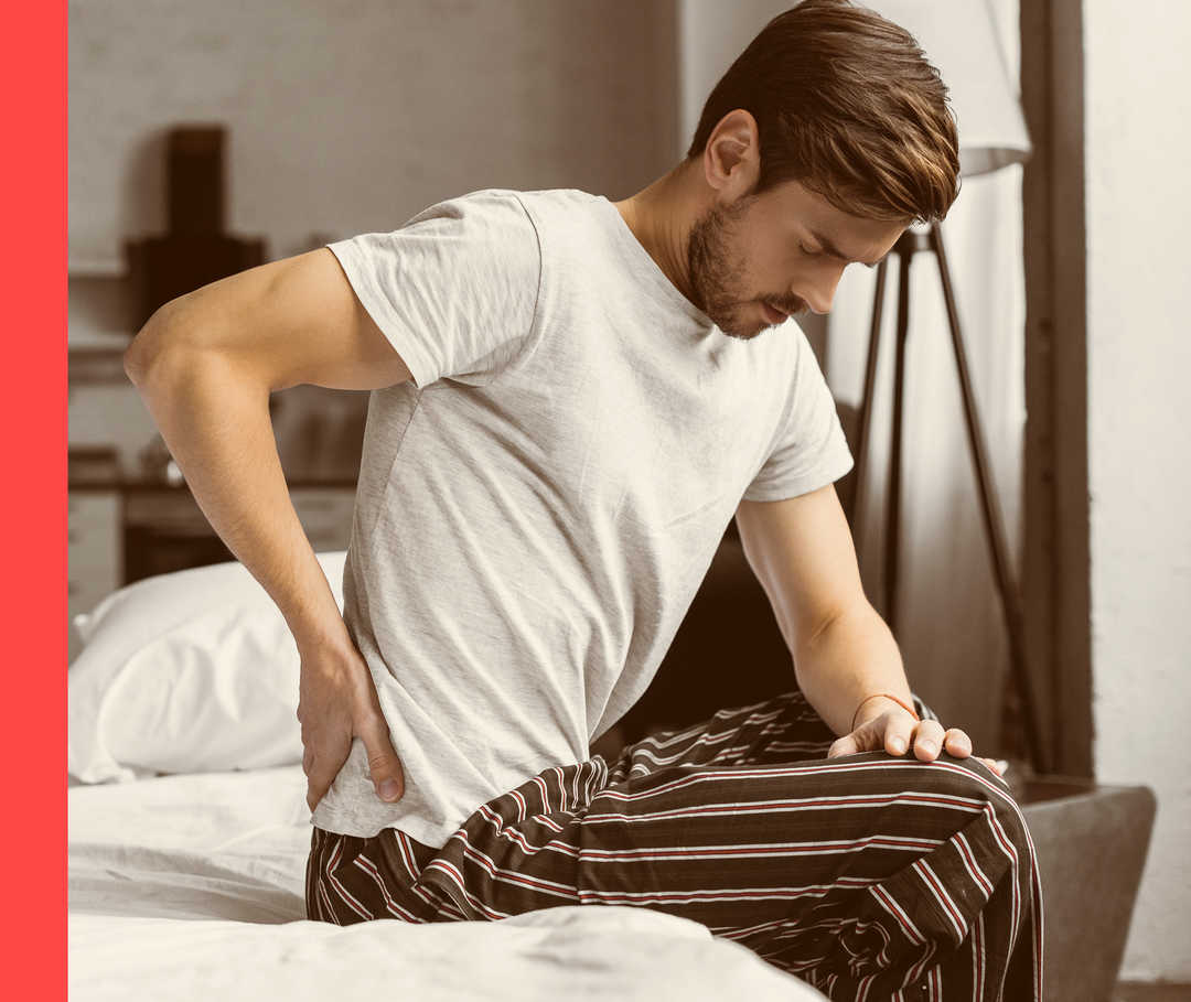 Mann sitzt im weißen T-Shirt und Schlafanzughose auf dem Bett und hält die Hand an seine rechte Rückenseite.
