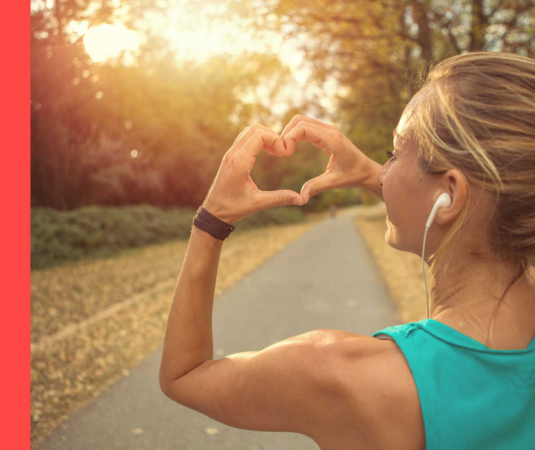 Ein Frau mit In-Ear-Kopfhörern und in einem türkisen Unterhemd hält ihre Finger in Herzform der Sonne entgegen.
